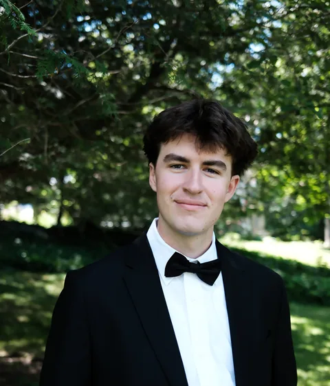 Portrait of Vincent Martino, smiling, in a dark jacket and bow tie, standing outdoors under trees
